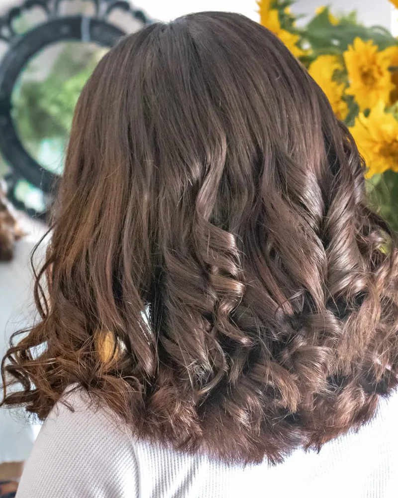 Cabelo castanho com caracóis largos e brilho, visto de costas, com girassóis ao fundo.