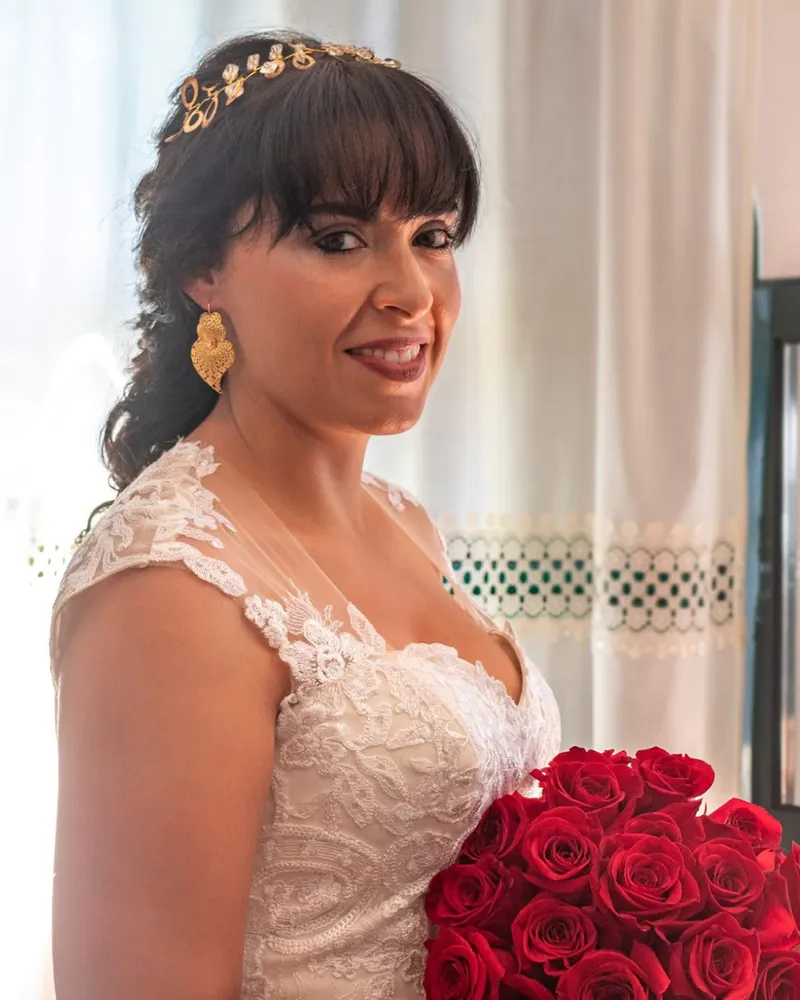 Noiva sorridente com franja, cabelo preso para trás e um acessório dourado, segurando um ramo de rosas vermelhas.